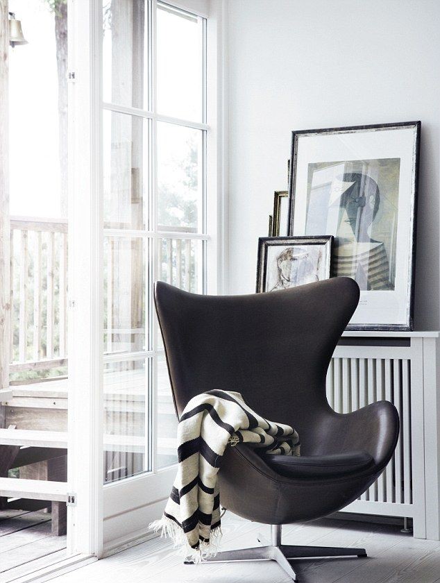 Arne Jacobsen - Chaise Egg en cuir noir sur un pied m&eacute;tal chrom&eacute;. Il est dans un angle d'une pi&egrave;ce blanche avec des cadres derri&egrave;res et une baie vitr&eacute;e ouverte. Une couverture ray&eacute;e blanche et noire avec des franges est pos&eacute;e sur le fauteil.